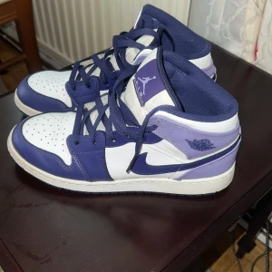 Nike Air Jordan 1 Mid Lila/Blå EUR 40 - Snygga Nike Air Jordan 1 Mid sneakers i vitt, lila och mörkblått skinn. Klassisk siluett med perforerad tå, ikoniska Jordan Wings-loggan och matchande blå snörning. Perfekta för dig som vill sticka ut med stilrena och trendiga sneakers.