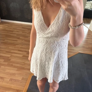 Vit spetsklänning med omlott - Supersöt vit kortklänning med omlott och v-ringad hals. har ett fint spetsmönster över hela tyget. Perfekt för sommarens alla tillfällen och studentfirande!