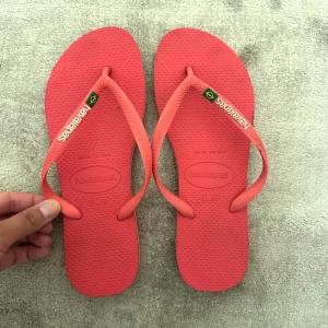 Rosa/korall flipflops från Havaianas - Säljer ett par snygga rosa flipflops från Havaianas med klassisk logga och brasiliansk flagga på remmen. Sulan har en randig detalj i gult och grönt på sidan! 