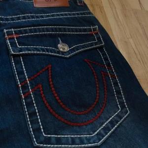 True Religion jeans med röd söm, stl 34 - Prislappen finns kvar och allt. Säljer ett par blå jeans från True Religion med ikoniska röda sömmar och kontrasterande vit stickning. Modellen har raka ben, stora fickor med lock och silverfärgade knappar. Jeansen är tillverkade i USA och har en cool streetstil som sticker ut.  