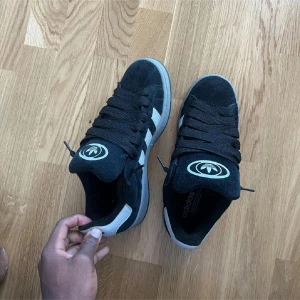 Adidas campus - Snygga svarta Adidas sneakers med klassiska vita ränder på sidorna och grå sula. Skorna har breda svarta skosnören och Adidas-logga på plösen och hälen. Ovandelen är i mocka och detaljer i syntet. Perfekta för en streetwear-look.