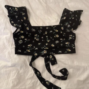 Svart blommig croppad topp med knyt - Superfin svart croppad topp med små vita och gula blommor. Toppen har volangärmar och knytband i midjan som ger en söt och trendig look. Perfekt för varma dagar och passar snyggt till både jeans och kjol.