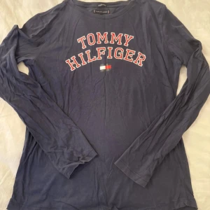 Marinblå långärmad Tommy Hilfiger tröja - Snygg marinblå långärmad tröja från Tommy Hilfiger med stort tryck i vitt och rött på bröstet. Tillverkad i mjuk ekologisk bomull, perfekt för dig som gillar klassisk stil med en modern touch. Rund hals och bekväm passform.