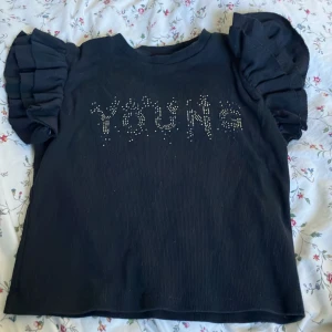 Svart topp med puffärmar från Gina Tricot - Svart topp från Gina Tricot med coola puffärmar och texten 'YOUNG' framtill i silvriga nitar. T-shirten har en rak passform och är tillverkad i ett mjukt, ribbat material. Perfekt för dig som vill sticka ut med en trendig och ungdomlig stil.