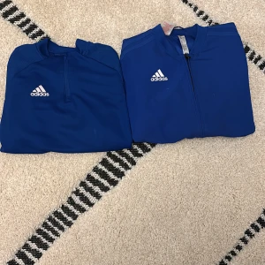 Blå adidas träningsjackor 140 - Två blå träningsjackor från adidas i storlek 140. Ena jackan har dragkedja hela vägen och den andra har half zip. Båda har klassisk adidas-logga på bröstet och är gjorda i ett lätt, sportigt polyestermaterial. Perfekta för träning eller chill.