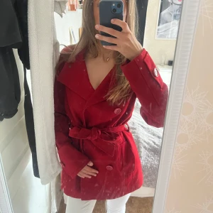 Röd kapp med knytning - Snygg röd trenchcoat med knytskärp i midjan. Jackan har dubbelknäppning med stora knappar och markerad midja.