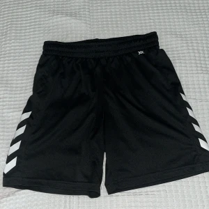 Svarta sportshorts från Hummel - Svarta sportiga shorts från Hummel med elastisk midja och vita detaljer i form av pilar på sidorna. Perfekta för träning eller fotboll, tillverkade i lätt och bekvämt polyestermaterial. Klassisk och stilren design med Hummel-logga på ena benet.