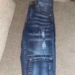 Blå Dsquared2 jeans  - Snygga blå jeans från Dsquared2 med coola slitningar och färgstänk på benen. Klassisk femficksmodell med Dsquared2-logga på fickan och bak. Jeansen har en modern passform och detaljer som ger en trendig look.