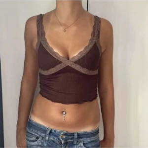 Brun ribbad croppad linnetopp - Säljer en brun croppad linnetopp med ribbad struktur och spetsdetaljer i beige längs kanterna. Toppen har breda axelband och omlott-design framtill som ger en snygg 90-talsvibe. Perfekt att styla med jeans eller kjol.