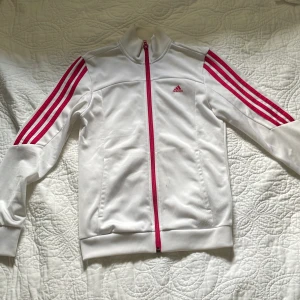 Vit Adidas vindjacka med rosa detaljer - Tröjan är i storlek 11-12 men jag som är xxs/xs kan ha den. Vit Adidas vindjacka med klassiska rosa ränder längs ärmarna och rosa dragkedja. Jackan har hög krage, två fickor och är tillverkad i polyester. Perfekt för dig som gillar sportig stil och vill sticka ut med färgade detaljer.