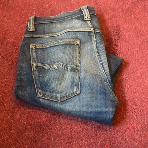 Blå jeans från Nudie Jeans, slimfit - Säljer ett par klassiska blåa slimfit jeans från Nudie. Modellen är grim tim, har fem fickor och Nudie-logga på bakfickan. Jeansen är i denim med en cool tvättad look och orange detaljer vid sömmarna. Det finns små defekter som visas på bild 4. Hör av er vid minsta fundering!  Mvh/Vasa begagnat🙌