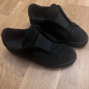 Axel Arigato Skor - Stilrena svarta sneakers från Axel Arigato i mocka med diskret logga på sidan och på plösen. Skorna har en platt sula och minimalistisk design utan snörning, vilket ger en clean look. Perfekta för dig som gillar enkel och modern stil. Använd fåtal gånger. Bra skick, pris kan diskuteras vid snabb affär