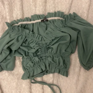 Grön omlott blus med snörning - Supersöt grön blus från Spanien med omlott-design och snörning framtill. Offshoulder-modell med långa puffiga ärmar och resår vid midjan och ärmsluten. Perfekt för dig som gillar trendiga och romantiska plagg.