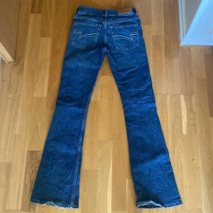 Blå bootcut jeans från Junkyard - Snygga blå jeans från Junkyard med bootcut passform och klassisk femficksdesign. Jeansen har coola bakfickor med vit zigzag-söm och ett brunt läderpatch med logga i midjan. Perfekta för dig som gillar retrovibbar och vill sticka ut lite extra.