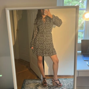 Svart blommig kortklänning med snörning - Supersöt svart kortklänning med vitt blommigt mönster och v-ringning med snörning i halsen. Klänningen har långa ärmar och ett lätt, luftigt tyg som ger en flowy känsla!!🫶🏻