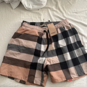 Burberry rutig badshorts med snörning - Snygga badshorts från Burberry med klassiskt rutigt mönster i beige, svart, vitt och rött. Modellen har elastisk midja med snörning och är tillverkad i lätt polyester som torkar snabbt. Perfekt för stranden eller poolen, riktigt stilrena och trendiga.