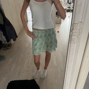 Grön blommig kortkjol med volang - Supersöt grön kortkjol med vitt blommigt mönster och volangkant. Har använt den  en gång bara så den har inte varit mycket använt alls