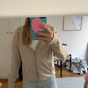 Beige hoodiejacka i kashmir från Soft Goat - Mysig beige hoodiejacka från Soft Goat i lyxig kashmir. Jackan har dragkedja, snörning i huvan och två fickor framtill. Perfekt för chill dagar och när du vill vara både bekväm och snygg. Långärmad och med ribbade muddar för extra komfort.