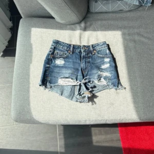 Blå slitna jeansshorts med fransar - Snygga blå jeansshorts från Levi's med hög midja, slitna detaljer och fransig kant. Klassisk femficksmodell med knapp och dragkedja. Perfekta för sommaren och ger en cool, avslappnad vibe.