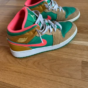 Nike Air Jordan 1 Mid SE Wheat Watermelon - Nike Air Jordan 1 Mid sneakers. Klassisk snörning, vit sula och ikonisk Jordan-logga i guld på sidan. 