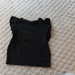 Svart topp med volangärm från 157 - Svart topp från 157 i mjuk bomullsblandning med elastan. Modellen har korta ärmar med volangdetalj som ger en söt och trendig look. Perfekt att matcha med jeans eller kjol för en chill stil.