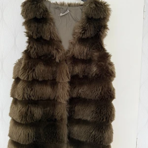 Brun fluffig pälsväst - Säljer en brun pälsväst med fluffiga, breda horisontella paneler. Västen har en öppen front och är helfodrad på insidan. Den har en loose passform och ger en riktigt cool och mysig vibe.