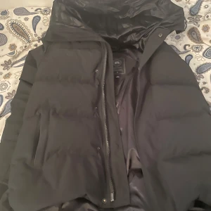 Svart pufferjacka från Canada Goose - Säljer en svart pufferjacka från Canada Goose med huva och snyggt märke på ärmen. Jackan har dragkedja framtill och två sidofickor. Den är vadderad och har en clean, stilren look som funkar perfekt när det är kallt ute.