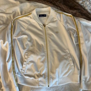 Vit Fred Perry bomberjacka med guld - Snygg vit sweatshirt från Fred Perry med guldfärgade ränder längs ärmarna och klassisk ribbad krage. Jackan har dragkedja framtill, två fickor och ikonisk Fred Perry-logga på bröstet. Perfekt för en sportig och stilren look.