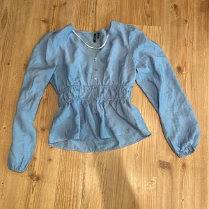 Blå blus med v-ringning från Vero Moda - Supersöt ljusblå blus från Vero Moda i storlek XS. Blusen har v-ringning, knappar framtill och markerad midja med resår. Långa ärmar med lätt puff och elastiska muddar. Perfekt för dig som gillar en feminin och trendig look.💞💞