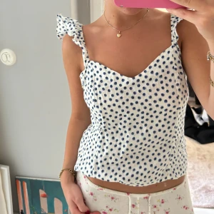 Prickig vit och blå linnetopp med volang - Supersöt vit linnetopp med blå prickar och volangdetaljer på axelbanden. Toppen har en lätt hjärtformad urringning och är ärmlös, perfekt för varma dagar🩷