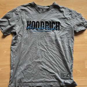 Grå t-shirt från Hoodrich med tryck - Snygg grå t-shirt från Hoodrich med svart och blå broderad logga framtill och tryck på ryggen. Klassisk passform med korta ärmar och rund hals. Patch med detaljer nertill. Perfekt för dig som gillar streetwear och vill sticka ut lite extra. Jag vill säja den här t-shirt för att jag inte använder den ofta och den ligger bara i garderoben.