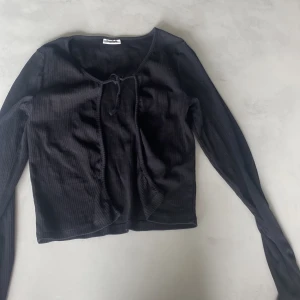 Svart ribbad kofta från Kappahl - Svart ribbad kofta från Kappahl med lång ärm och knytning framtill. Modellen är tunn och har en enkel, stilren look som passar perfekt till lager-på-lager. Materialet är mjukt och stretchigt för en bekväm känsla hela dagen.