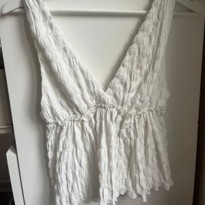 Vit topp  med v-ringning - Supersöt vit omlott linnetopp med djup v-ringning och breda axelband. Toppen har en luftig, vågig struktur och knytning i midjan som ger en avslappnad och somrig känsla. Perfekt till varma dagar och lätt att matcha med jeans eller kjol.