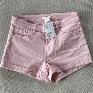 Ljusrosa shorts från H&M, strl 34 - Snygga ljusrosa shorts från H&M i klassisk jeansmodell med fem fickor och uppvikta benslut. Perfekta för varma dagar och lätta att matcha med allt. Gjorda i mjuk bomull med lite stretch för extra komfort. Passar dig som gillar en enkel och fräsch stil.