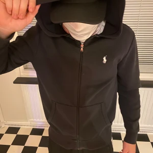 Svart hoodie från Ralph Lauren - Svart hoodie från Ralph Lauren med dragkedja och huva. Tröjan har två fickor framtill och den klassiska Polo-loggan broderad på bröstet. Tillverkad i mjuk bomull, perfekt för chill dagar. Långärmad och med ribbade muddar för extra komfort.