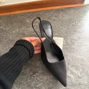 Svarta pumps med spetsig tå från H&M - Stilrena svarta pumps från H&M med spetsig tå och smal klack. Skorna har en elegant slingback-rem och är tillverkade i syntetmaterial med mockakänsla. Perfekta för dig som vill ha en clean och trendig look.