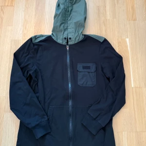 Diesel hoodie  - Svart Diesel hoodie med dragkedja och en olivgrön huva. Jackan har en ficka på bröstet och en etikett med texten "D78 ONLY THE BRAVE" nedtill. Storlek S