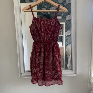 Vinröd kortklänning Abercrombie & Fitch - Supersöt vinröd kortklänning från Abercrombie & Fitch med tunna axelband och ett guldigt blommigt mönster. Klänningen har en markerad midja med resår och är tillverkad i ett lätt och luftigt material som passar perfekt när du vill känna dig extra fin.