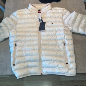 Vit pufferjacka från Tommy Hilfiger - Snygg vit pufferjacka från Tommy Hilfiger med diskret logga på bröstet och dragkedjor på fickorna. Grym höstjacka, endast använd 2 gånger. Prislapp sitter kvar. Köpt på en original Hilfiger butik i Barcelona, därav euro på prislappen. Skick 10/10, i princip helt ny.