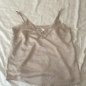 Beige linnetopp med spets från Kappahl - Supersöt beige linnetopp från Kappahl med tunna axelband och v-ringning dekorerad med blommig spets. Sidenliknande material som känns lyxigt och lätt. Perfekt att styla med jeans eller kjol för en trendig look.