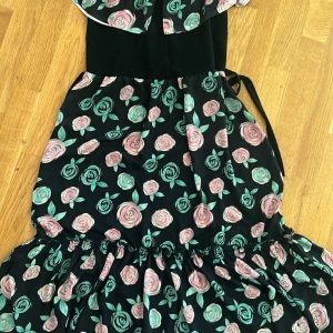 Svart långklänning med blommigt mönster - Säljer en svart långklänning med rosa och gröna blommor. Klänningen har volangdetaljer och knytband i midjan. Materialet känns lätt och flowy, perfekt för dig som gillar romantiska vibes och färgglada prints.