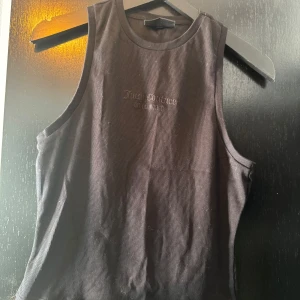 Svart ribbad linnetopp Juicy Couture - Svart ribbad linnetopp från Juicy Couture med broderad logga framtill. Toppen är ärmlös och har en klassisk rund halsringning. Perfekt för en clean och stilren look, lätt att matcha med både jeans och kjol.