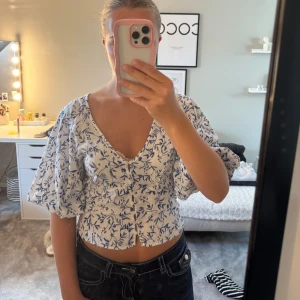 Vit och blå blommig blus med puffärm - Superfin croppad blus från H&M i vit med blått blommönster. V-ringad med knappar framtill och stora puffärmar. Luftigt material.💕