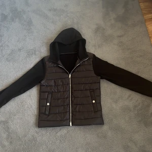 Svart pufferjacka med huva från H&M - Svart cardigan  från H&M i storlek S. Jackan har quiltad framsida, dragkedja, två fickor med tryckknappar och en skön huva. Ärmar och baksida är i mjuk bomull, medan framsidan är vadderad och i polyester. Perfekt för kyliga dagar. Den är helt ny och den är stor på mig 
