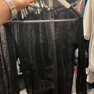 Svart mesh med lång ärm - Säljer en svart, transparent mesh tröja med långa ärmar. Toppen har en lösare passform och är perfekt för att styla lager på lager. Tunn och stretchig med diskret volangkant vid halsen. Superenkel att matcha till olika outfits. 