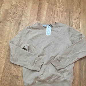 Beige sweatshirt från 157, storlek 150cl - Säljer en stilren beige sweatshirt från 157 i storlek 150cl. Tröjan har rund hals, långa ärmar och ribbade muddar vid ärmslut och nederkant. Perfekt för dig som gillar enkel och clean stil. Materialet är mjukt och skönt, troligtvis bomull eller polyester.