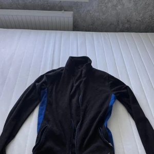 Svart fleecejacka från Bluewear - Svart fleecejacka från Bluewear med blåa paneler längs sidorna. Jackan har hög krage, långa ärmar och dragkedja framtill samt vid fickorna. Vid frågär hör av dig!