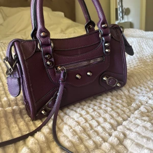 Lila axelväska i skinn med detaljer - Snygg lila axelväska i skinn med silverfärgade metalldetaljer och dragkedja framtill. Väskan har både handtag och en lång axelrem, samt coola nitar och spännen som ger en edgy vibe. Perfekt storlek för det viktigaste och har ett rymligt fack med dragkedja.