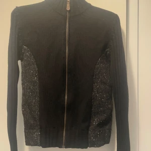 Svart kofta med dragkedja från Hugo Boss - Svart kofta från Hugo Boss med snygga stickade paneler och grov dragkedja framtill. Tröjan har långärmade ribbade ärmar och detaljer i mörkgrått på sidorna. Materialet är 100% ull vilket gör den både varm och mjuk. Perfekt för dig som gillar stilrena och klassiska plagg. Använd ca 2-3 gånger