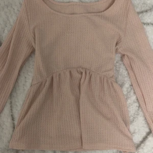 Ljusrosa oanvänd långärmad blus - Säljer en söt ljusrosa blus med lång ärm och rundad hals. Blusen har en peplumformad midja och är tillverkad i ett mjukt, strukturerat bomullstyg. Perfekt för dig som gillar romantisk och feminin stil.